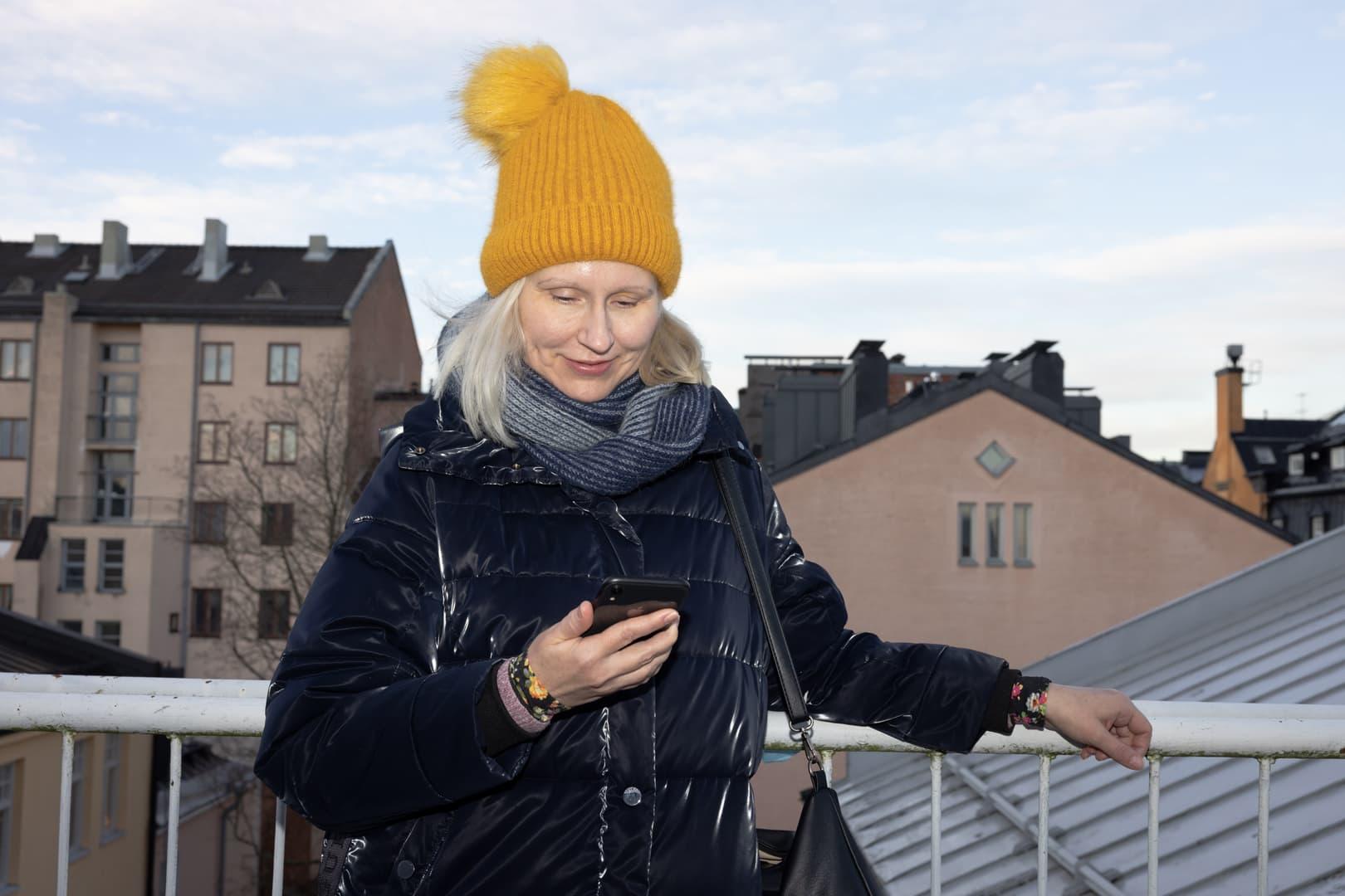 En Anyfinkund står på sin balkong och tittar ner i sin mobil. Bakom henne ser man takåsar.