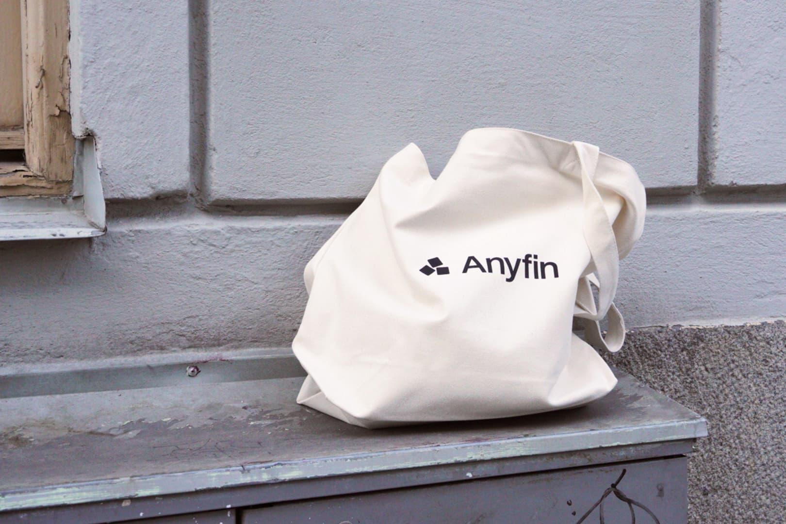 Kuvassa on Anyfin logolla varustettu valkoinen kangaskassi, joka on asetettu talon seinälle olevalle ikkunalaudalle