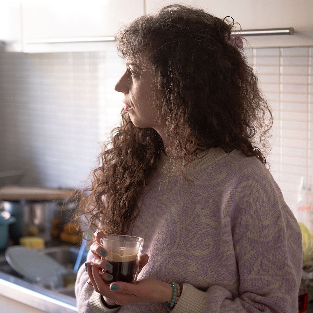 En kvinnar står i sitt kök med en kaffekopp i handen.