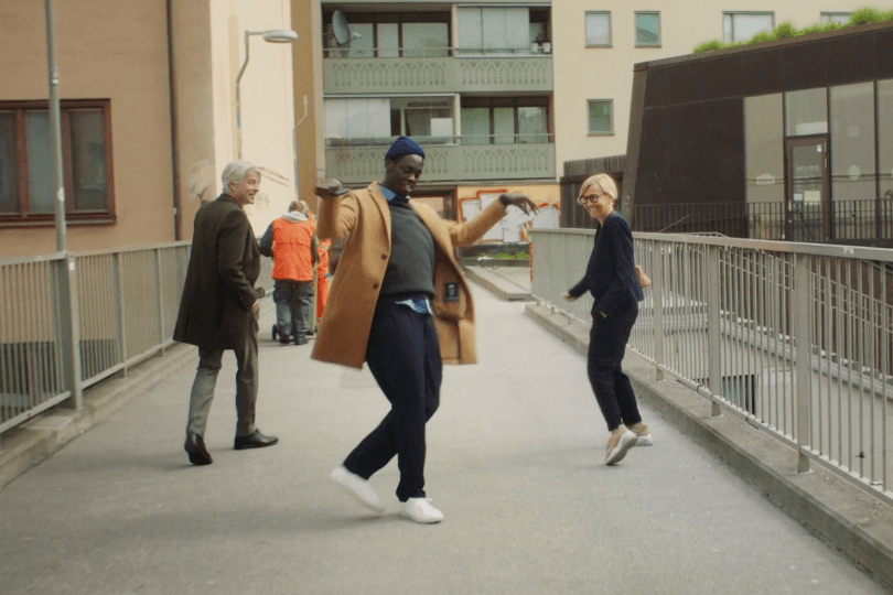 Man in beige coat dancing on pedestrian bridge with two women looking at him and laughing