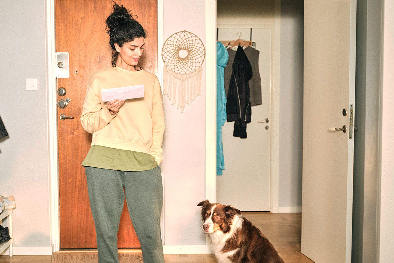 Anyfinkund som står i en hall med en elräkning i ena handen och tittar ner på hunden som sitter bredvid.