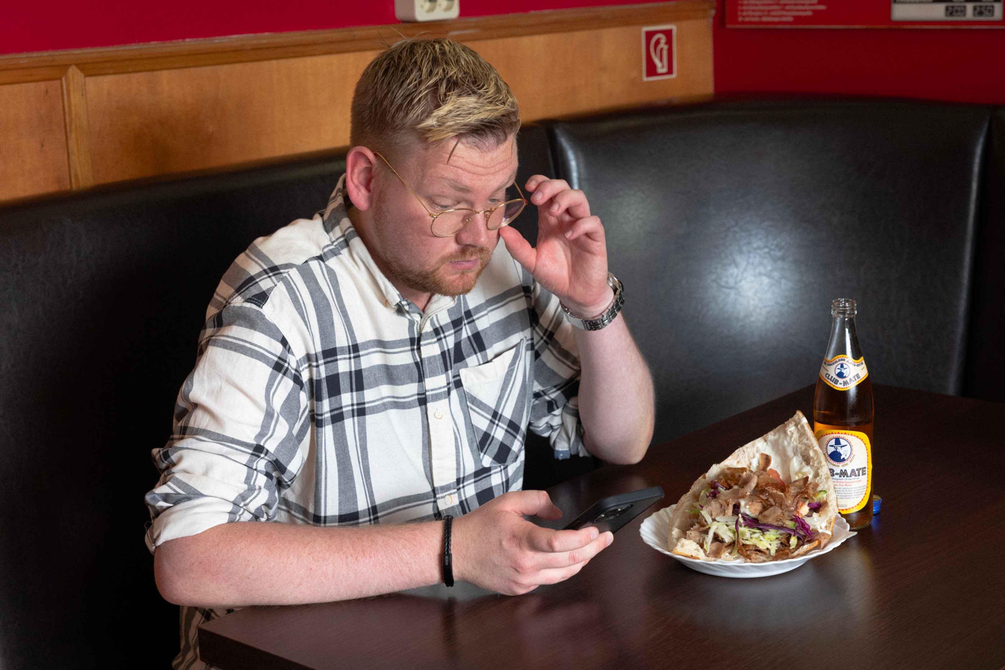 Man som sitter på en restaurang och tittar på mobilen. Framför sig har han en kebab i bröd.